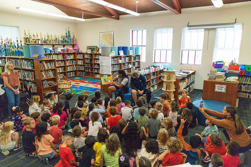 A group of students sit on the floor of the library listening to two college football players reading to them