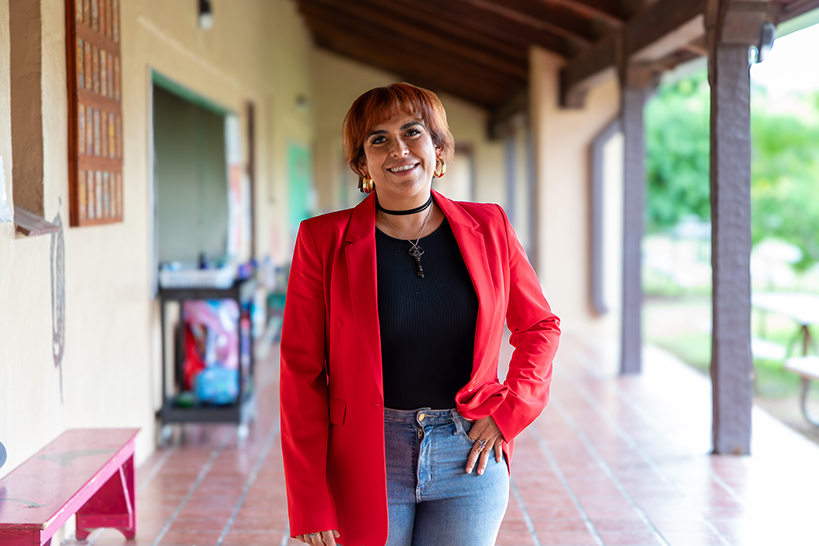 A woman in a red blazer and jeans smiles outside the school